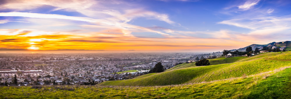 Sunset view of residential and industrial areas in East San Francisco Bay Area; green hills visible in the foreground; Hayward, California
